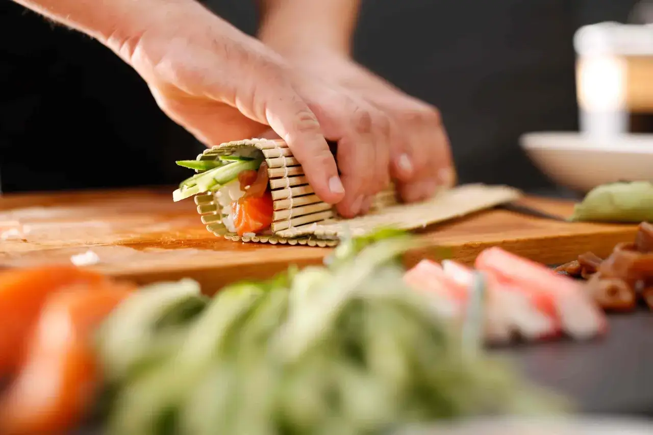 Ręce zwijają sushi z łososiem i ogórkiem, posypane sezamem.