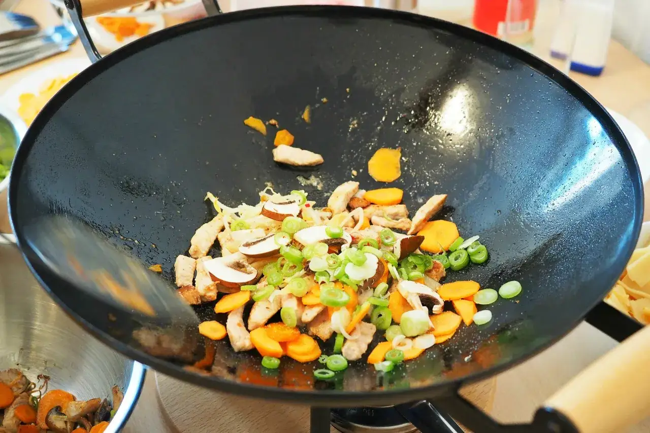 Na patelni wok smażą się kawałki kurczaka, marchewka, pieczarki i szczypiorek. Idealne składniki do szybkich wok przepis&oacute;w.