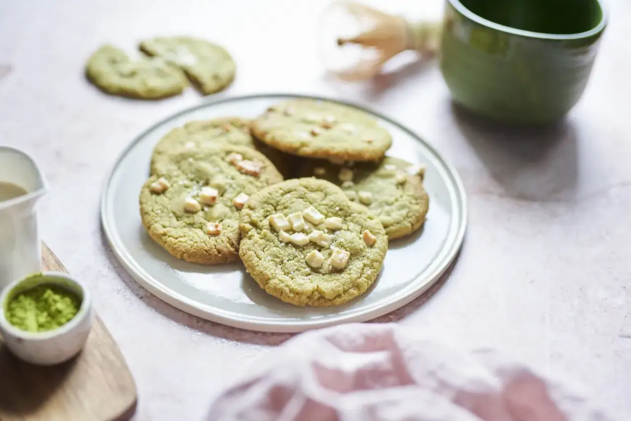 Zielone ciasteczka matcha z kawałkami białej czekolady na talerzu, obok miseczki z matchą i mlecznika.