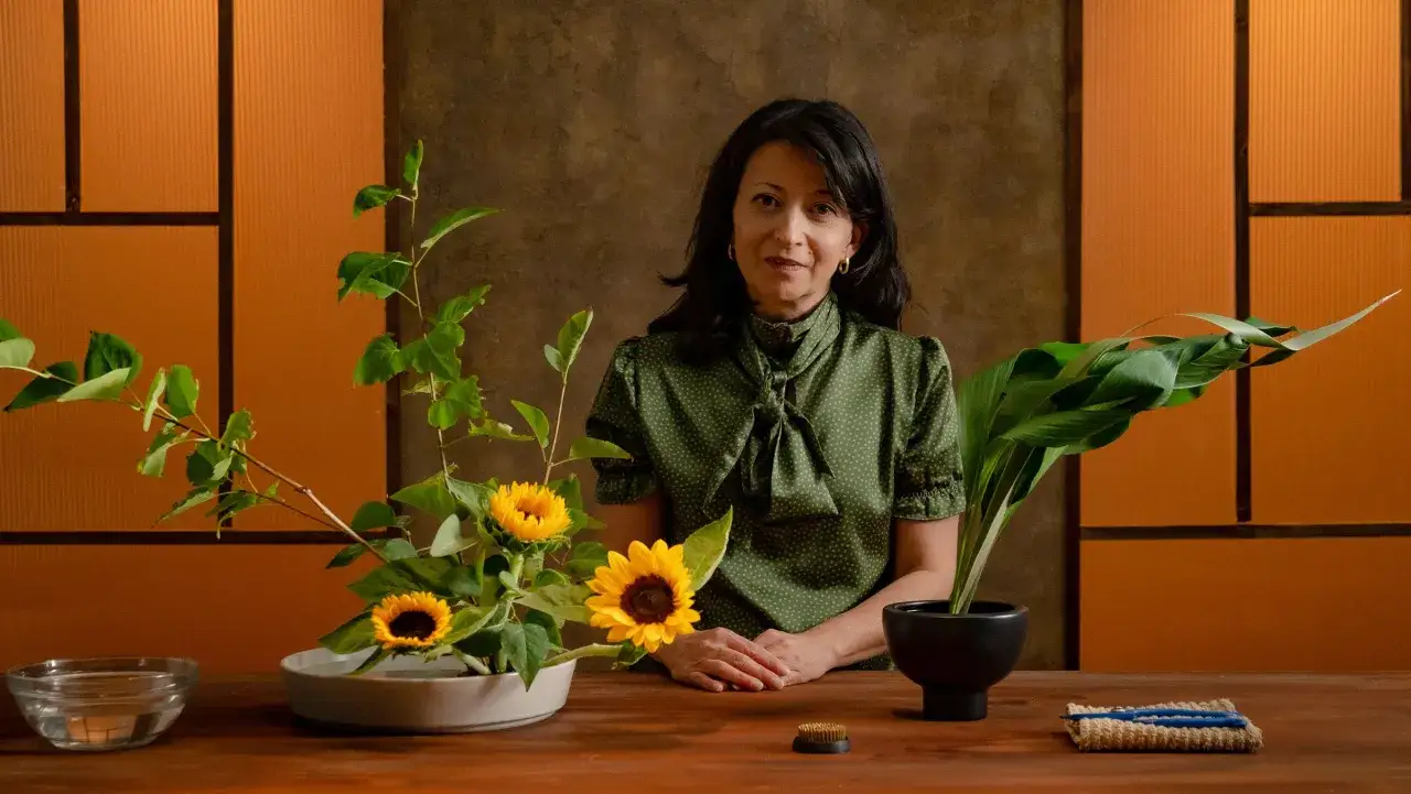 Kobieta w zielonej bluzce prezentuje dwie kompozycje ikebana: słoneczniki i liście.