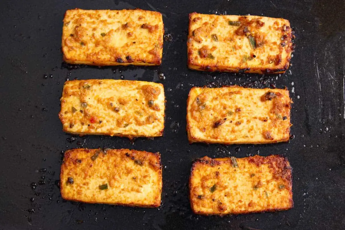 Sześć kawałków smażonego miso tofu, lekko przypieczonych, z zielonymi dodatkami, na czarnej patelni.