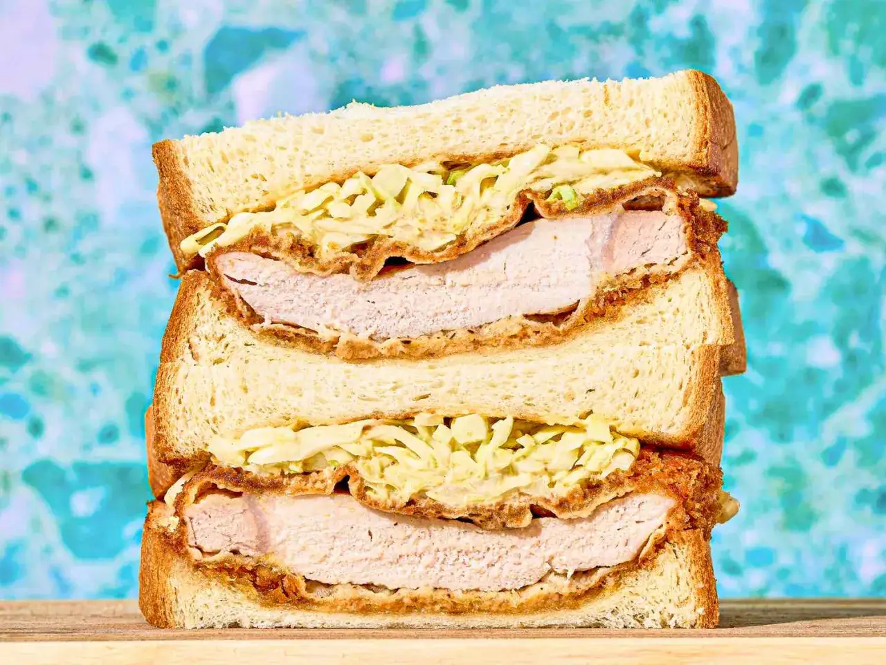 Dwa piętrowe katsu sando z chrupiącym kotletem i surówką na tle turkusowej ściany.