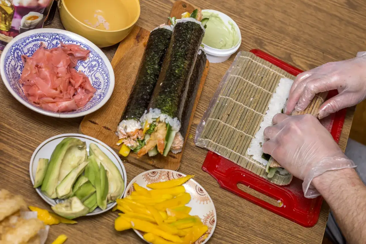 Przygotowanie sushi: ryż, awokado, mango, krewetki i marynowany imbir. Ręce w rękawiczkach zwijają rolkę na macie bambusowej. Jaka sałata do sushi? Tutaj jej nie widać, ale składniki są idealne!