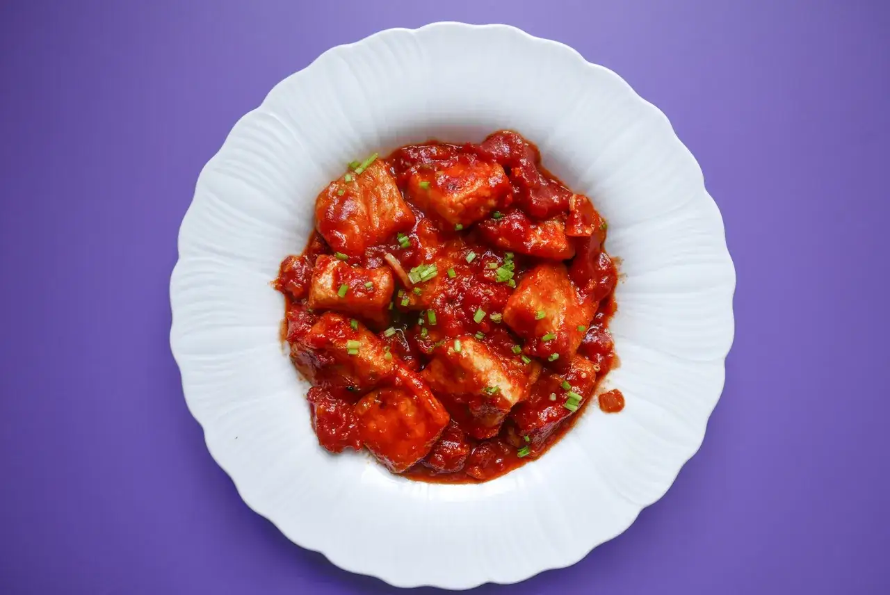Kawałki tofu w sosie pomidorowym, posypane szczypiorkiem, na białym talerzu.