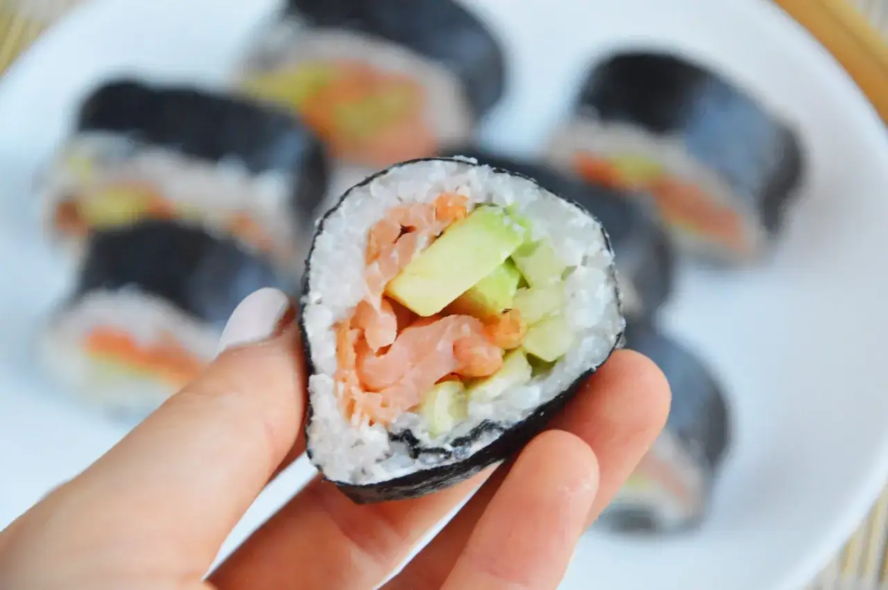 Smaczne sushi z łososiem, awokado i og&oacute;rkiem, trzymane w dłoni.