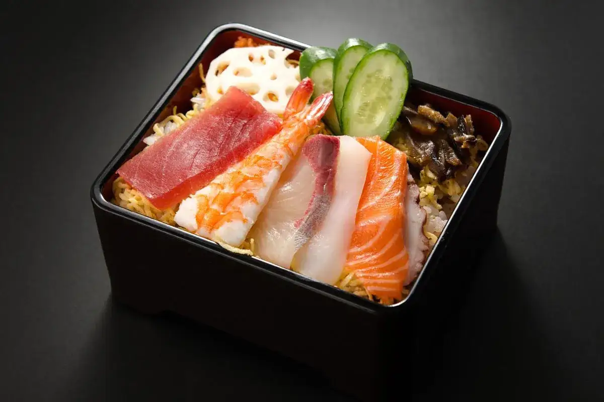 Chirashi zushi: kolorowe plastry ryb (łosoś, tuńczyk, biała ryba), krewetka, plasterki og&oacute;rka i kwiat lotosu na ryżu.