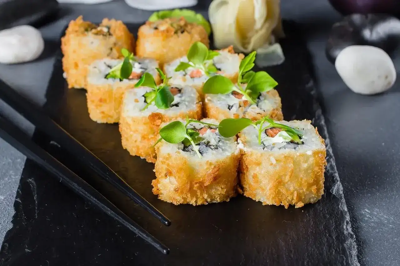 Chrupiące rolki sushi, w tym california roll, podane na czarnej desce z pałeczkami i zielonymi kiełkami.