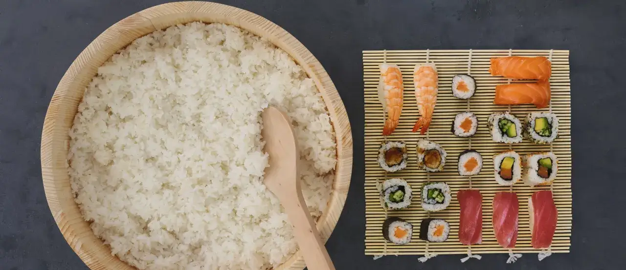 Gotowy ryż do sushi w drewnianej kadzi, obok mata bambusowa z różnymi rodzajami sushi. Zastanawiasz się, ile octu do sushi?