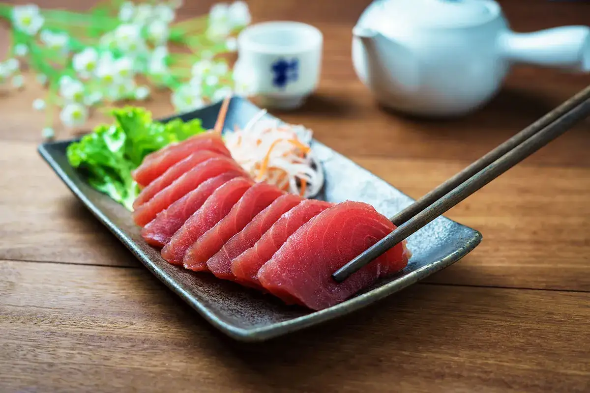 Sashimi z tuńczyka, czyli wykwintna ryba japońska, prezentowana na talerzu z sałatą i startymi warzywami. Pałeczki gotowe do degustacji.